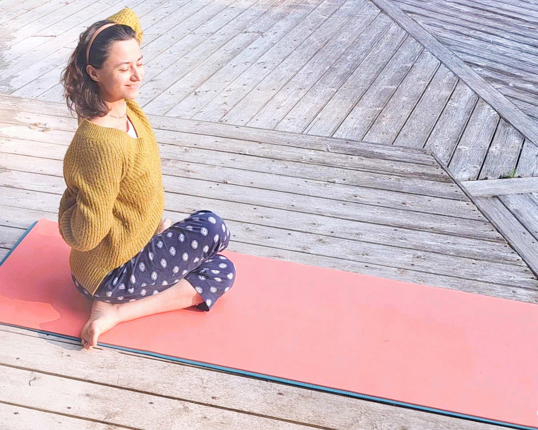Zofia sitting cross-legged on a coral yoga mat on a sunlit wooden deck